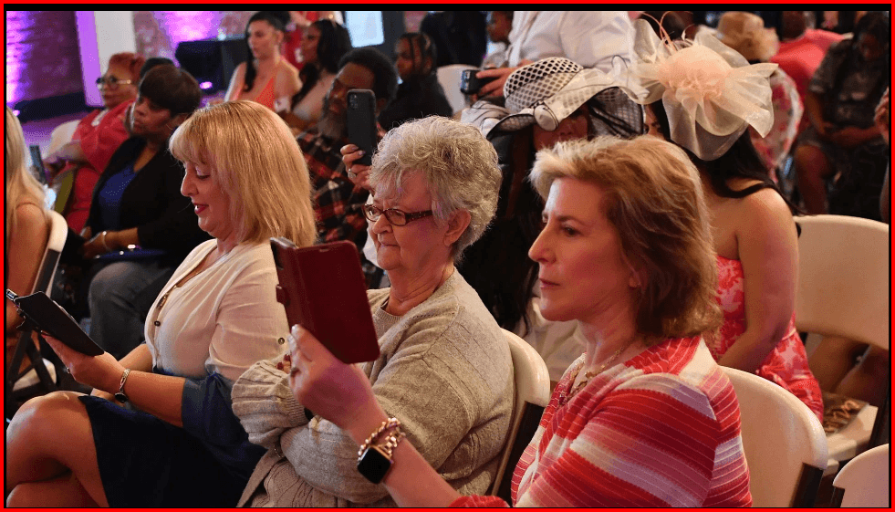 Group of women seated in an audience, some holding smartphones to capture the event.